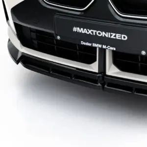 Close-up of a black car front with a license plate displaying #MAXTONIZED and BMW emblem nearby on a light background.