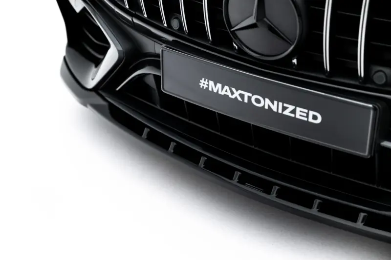 pol_po_Set-of-Splitters-Mercedes-AMG-C63-Sedan-Estate-W206-S206-23828_12webp - OPSHOLDERS Close-up of a glossy black car grille with chrome vertical bars and a plate showing #MAXTONIZED.