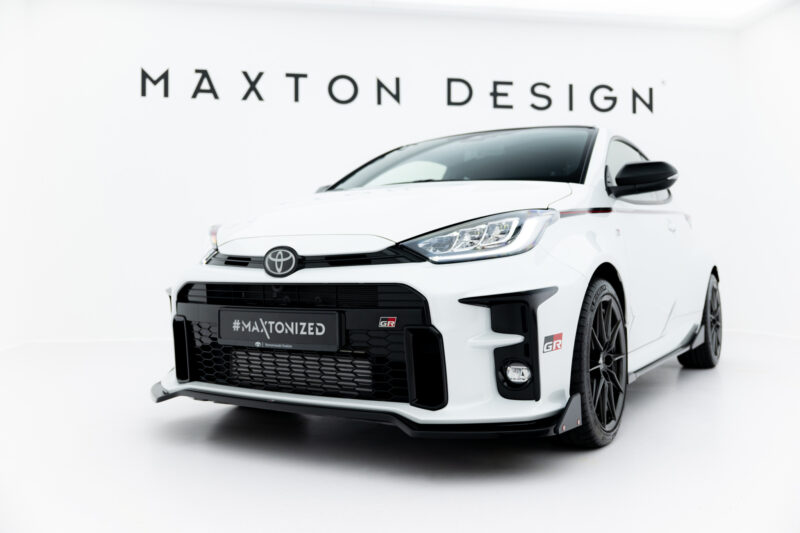 White Toyota sports car photographed from a low front-right angle in a bright showroom; background shows the brand 'MAIXTON DESIGN' (Maxton Design) sign and the car's grille and headlights are prominent. The license plate reads '#MAXTONIZED'.