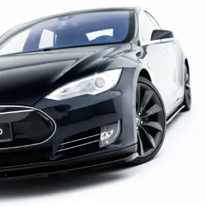 Close-up of a black Tesla car's front-left area, showing the headlight, emblem, and glossy body against a white background.