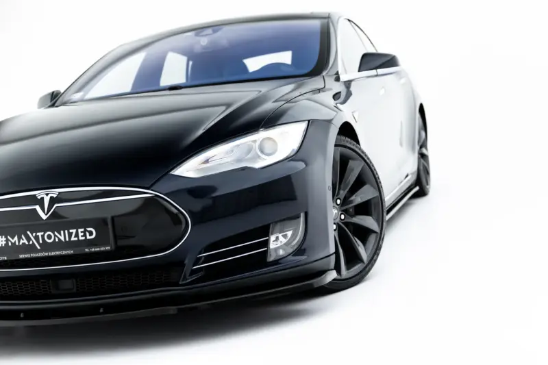 Close-up of a black Tesla car's front-left area, showing the headlight, emblem, and glossy body against a white background.