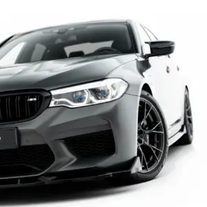 Front-left angle of a dark gray BMW M5, showing the grille, headlight, and front wheel with a #MAXTONIZED plate tag visible on the bumper.