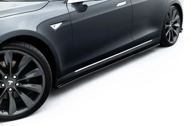 Side view of a dark gray Tesla sedan, showing the front wheel, door handle, and smooth body lines with a visible Tesla logo badge.