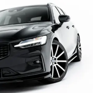 Front-left quarter view of a glossy black Volvo car with a prominent grille, LED headlight, and large multi-spoke alloy wheel against a white background.