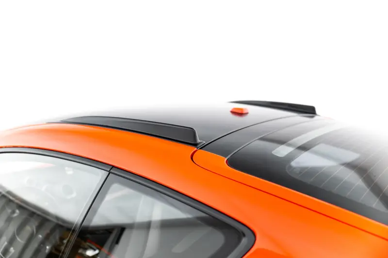 Close-up of an orange car roof with a black panoramic panel and small orange roof antenna, showing roof rails and curved windshield edges.