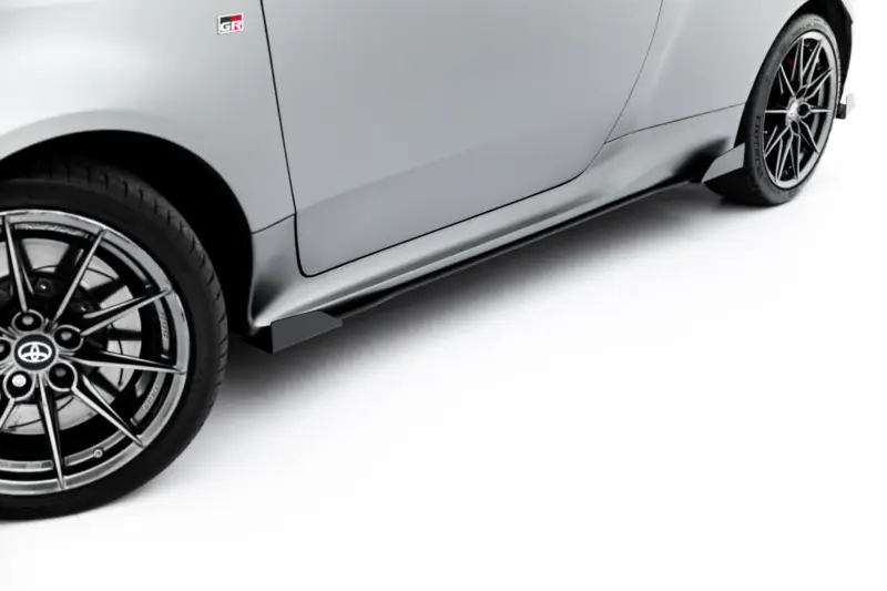 Close-up of a grey sports car's front wheel and side panel, displaying alloy wheel, low-profile tire, and a red brake caliper behind sleek spokes (GR badge visible).