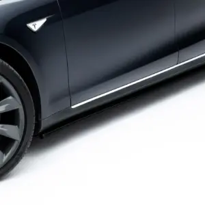 Close-up of a dark Tesla car's front passenger-side wheel and door panel, showing the Tesla emblem on the fender and chrome trim along the bottom edge