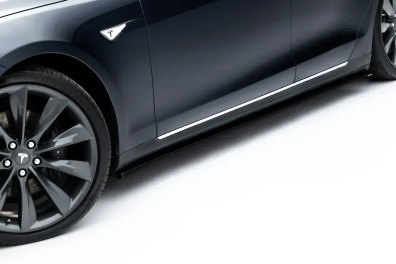 pol_po_Side-Skirts-Diffusers-V-1-Tesla-Model-S-Mk1-23946_2webp - OPSHOLDERS Close-up of a dark Tesla car's front passenger-side wheel and door panel, showing the Tesla emblem on the fender and chrome trim along the bottom edge