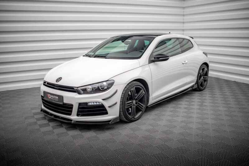 White two-door sports car parked indoors in front of a light gray, corrugated wall and checkered metal floor; visible VW badge on the hood.