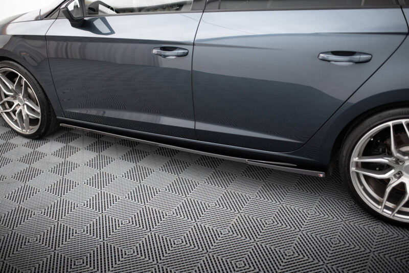Close-up of a gray car's side with two doors, chrome handles, a side mirror, and alloy wheels on a patterned floor.