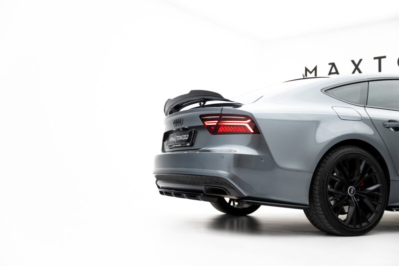 Rear-right view of a silver Audi with the trunk open and red taillights illuminated in a bright showroom; partial wall text visible in the background (MAX...).