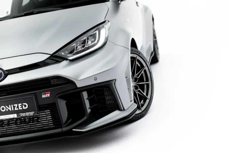 Close-up of a silver car's front-left corner, showing the headlight, bumper, and alloy wheel on a white backdrop.