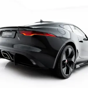 Rear three-quarter view of a dark Jaguar sports car with distinctive red tail lights and dual exhausts in a studio setting behind a white background.