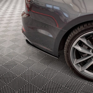 Rear quarter of a dark gray Audi, showing the rear bumper, wheel, and red brake calipers on a checkered floor in a showroom-like setting.