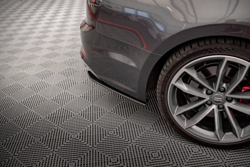 Rear quarter of a dark gray Audi, showing the rear bumper, wheel, and red brake calipers on a checkered floor in a showroom-like setting.