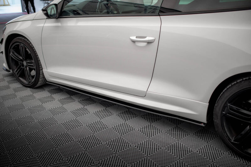 White car parked indoors; side door, handle, and front wheel with a black alloy rim visible in a showroom setting.