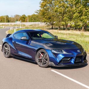 Blue sports car with a carbon-fiber hood parked on a sunny road, trees in the background.