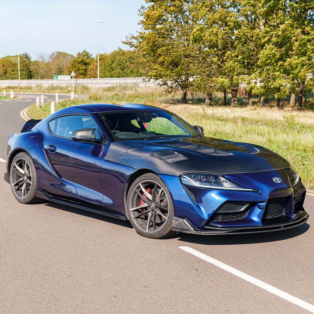 A90SupraV2Kit__OriginalCrop_CTCarbon_190925_30jpg - OPSHOLDERS Blue sports car with a carbon-fiber hood parked on a sunny road, trees in the background.