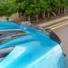 Artura_Web_CT_Carbon_170426_09jpg - OPSHOLDERS Blue sports car hood with a black carbon-fiber rear spoiler, parked near a hedge-lined curb.
