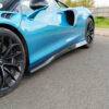 Close-up of a blue sports car's side; prominent air intake, carbon fiber side skirt, and large black alloy wheel visible.