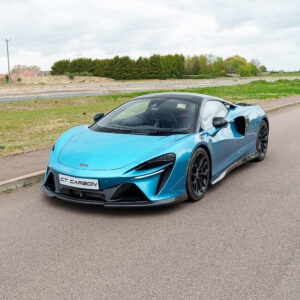 Blue McLaren-style sports car parked on a suburban road, front three-quarters view with green hedges in the background.