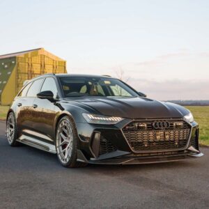 Front three-quarter view of a black Audi sports car with a large honeycomb grille and wide low-profile tires parked on a paved road at sunset/nearby industrial backdrop.