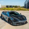 DSC05211-Enhanced-NR_e9068420-034c-4471-a50c-af39a95016d9jpg - OPSHOLDERS Blue-gray McLaren sports car with carbon fiber panels parked on a concrete lot, industrial buildings in the background, yellow brake calipers visible