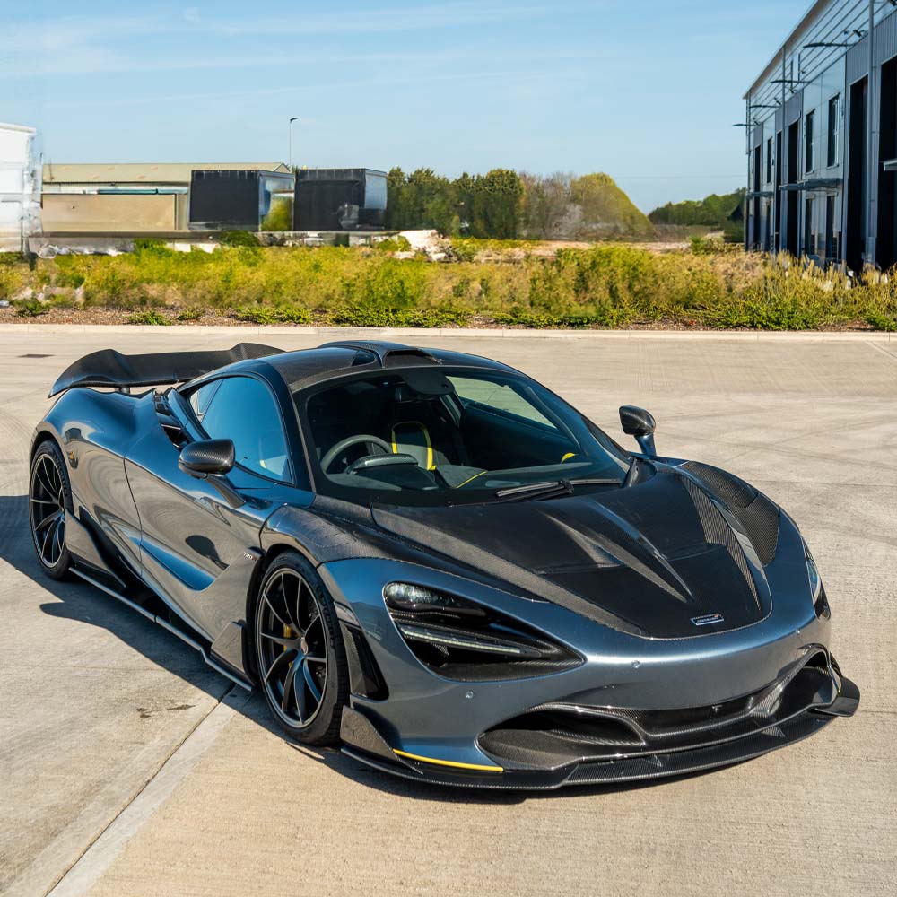DSC05211-Enhanced-NR_e9068420-034c-4471-a50c-af39a95016d9jpg - OPSHOLDERS Blue-gray McLaren sports car with carbon fiber panels parked on a concrete lot, industrial buildings in the background, yellow brake calipers visible