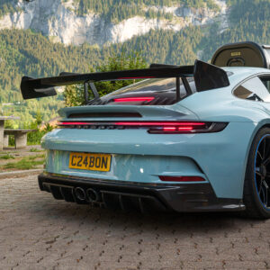 Rear view of a light-blue Porsche with a large rear wing and quad exhausts, set against a mountainous backdrop, license plate C24BON.