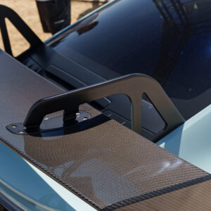 Close-up of a carbon fiber roof panel on a blue sports car, showing a black mounting bracket or handle. Inset bolts visible for added detail.