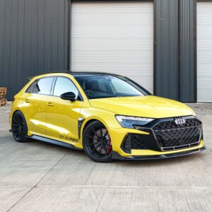 Yellow modified Audi sedan with black large-mesh front grille, carbon front splitter, and black aftermarket wheels parked in front of a dark industrial building