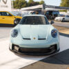Front view of a light blue Porsche sports car at an outdoor car show, with Monaco banners and other vehicles in the background