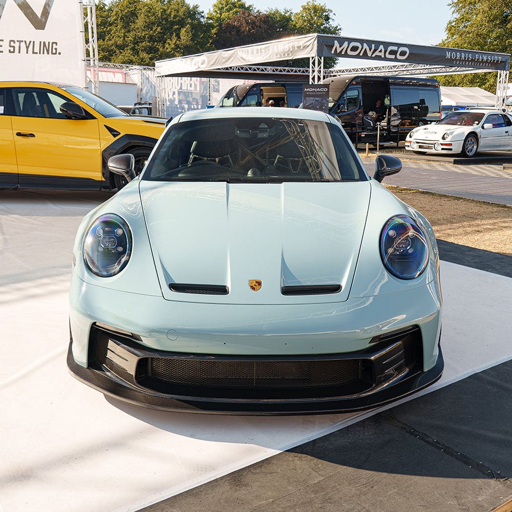GoodwoodFOS26_GT3_OriginalCrop_CTCarbon_100726_20copy_2e2b9fc5-7c1f-4301-9c6e-08a9e61e5472jpg - OPSHOLDERS Front view of a light blue Porsche sports car at an outdoor car show, with Monaco banners and other vehicles in the background