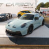 Light blue Porsche 911 on display platform at a car show, with another sleek car and people in the background under a large banner.