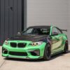 IMG_2290jpg - OPSHOLDERS Front three-quarter view of a lime-green BMW sports car with a carbon fiber hood and black roof, parked beside a grey industrial building.