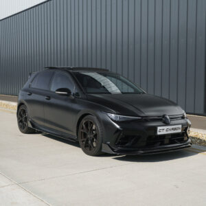 Black VW hatchback with carbon aero kit parked beside a gray industrial building; sleek, sporty appearance.
