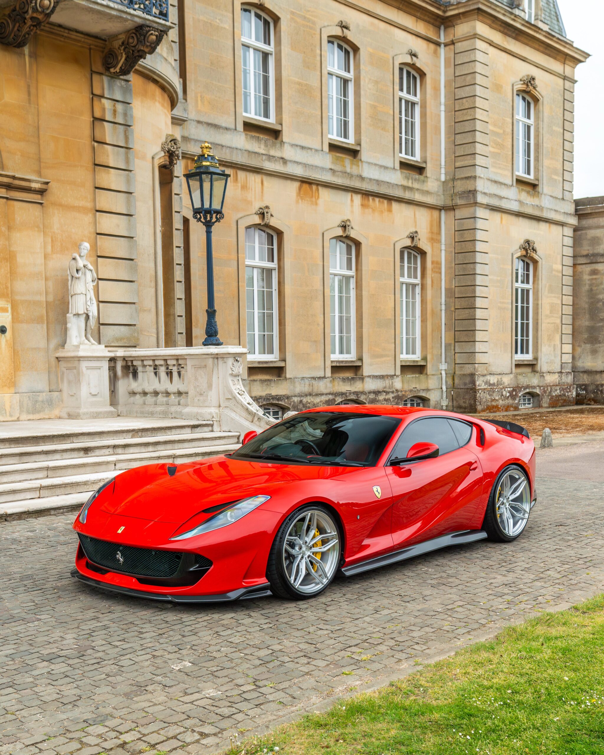 Red Ferrari sports car parked on a cobblestone driveway beside a statue and ornate blue street lamp in front of a beige historic stone building.](https://example)