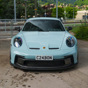 Front view of a light-blue Porsche 911 with a black front splitter and rain droplets on the windshield, license plate C24BON.