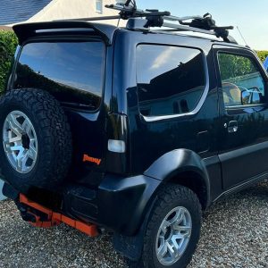 Rear view of a black compact SUV with a spare tire mounted on the back and a roof rack on top, parked on gravel.
