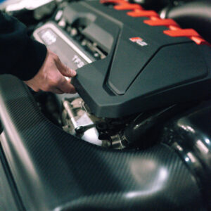 25-tfsi-engine-cover-grommets_53352784778_ojpg - OPSHOLDERS A mechanic's hand grips a panel of a car engine, exposing the engine bay beneath with red accents on the cover.