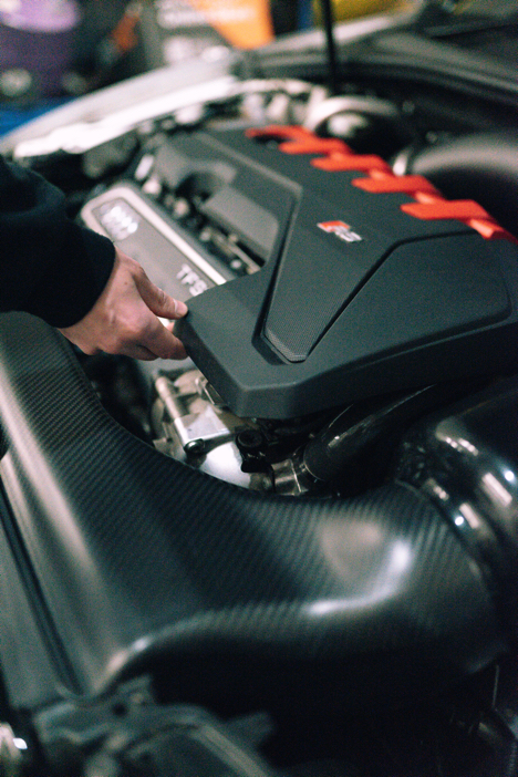 25-tfsi-engine-cover-grommets_53352784778_ojpg - OPSHOLDERS A mechanic's hand grips a panel of a car engine, exposing the engine bay beneath with red accents on the cover.
