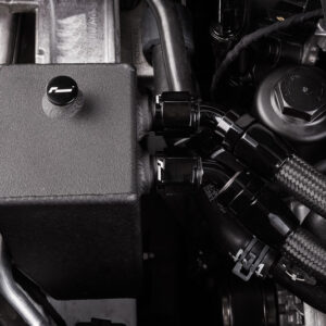 Close-up of a car engine bay featuring a black rectangular reservoir and thick braided hoses and clamps for connection.