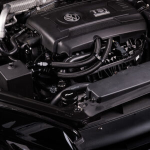 Close-up of a car engine bay with a Volkswagen engine cover and surrounding hoses and components at the front of the vehicle.