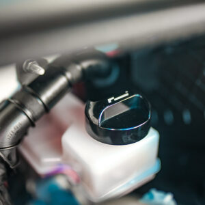 50589894726_2fd28d1161_cjpg - OPSHOLDERS Close-up of a white plastic reservoir with a black cap inside a car's engine bay.