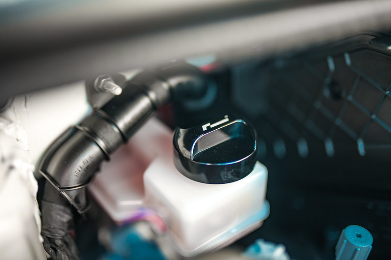 50589894726_2fd28d1161_cjpg - OPSHOLDERS Close-up of a white plastic reservoir with a black cap inside a car's engine bay.