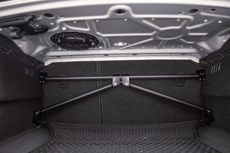 51286203405_53812351a2_k-1jpg - OPSHOLDERS Interior of a car trunk viewed from below, showing the underside of the trunk lid with structural crossbars, a circular vent/grill, and bundled support rods against dark carpeted walls