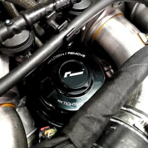 Close-up of a black oil cap on a motorcycle engine, showing 'Remove' and 'Tighten' markings, surrounded by hoses and metal pipes.