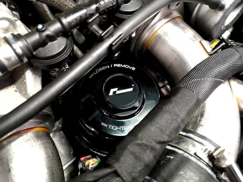 Close-up of a black oil cap on a motorcycle engine, showing 'Remove' and 'Tighten' markings, surrounded by hoses and metal pipes.