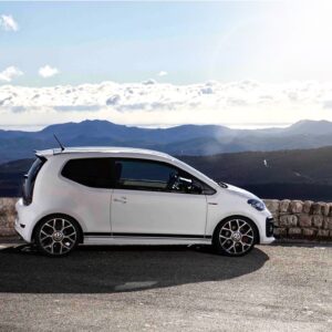 White compact car parked along a stone barrier with a mountain range and blue sky in the background.