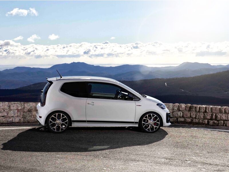White compact car parked along a stone barrier with a mountain range and blue sky in the background.